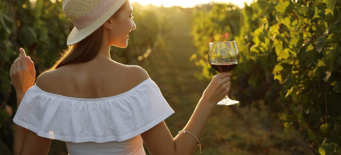 Beautiful young woman with glass of wine in vineyard on sunny day, back view