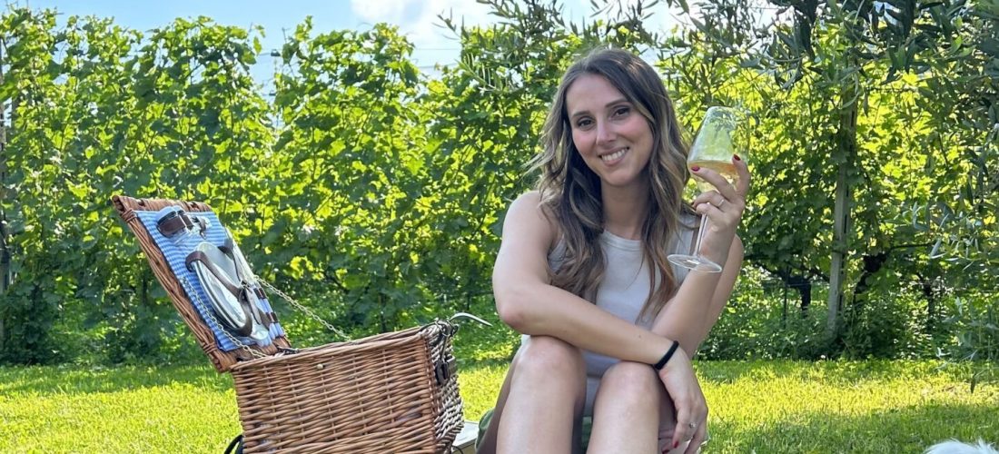 Ragazza seduta su telo con cestino da picnic sotto alberi di fronte a vigneti bresciani in Val Camonica